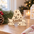 Houten kerstbalhanger in de vorm van een grote letter A met sterren en naam Amelia in een banner.